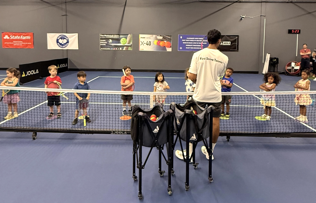 Young kids lined up on an indoor tennis court with a First Serve Tennis coach